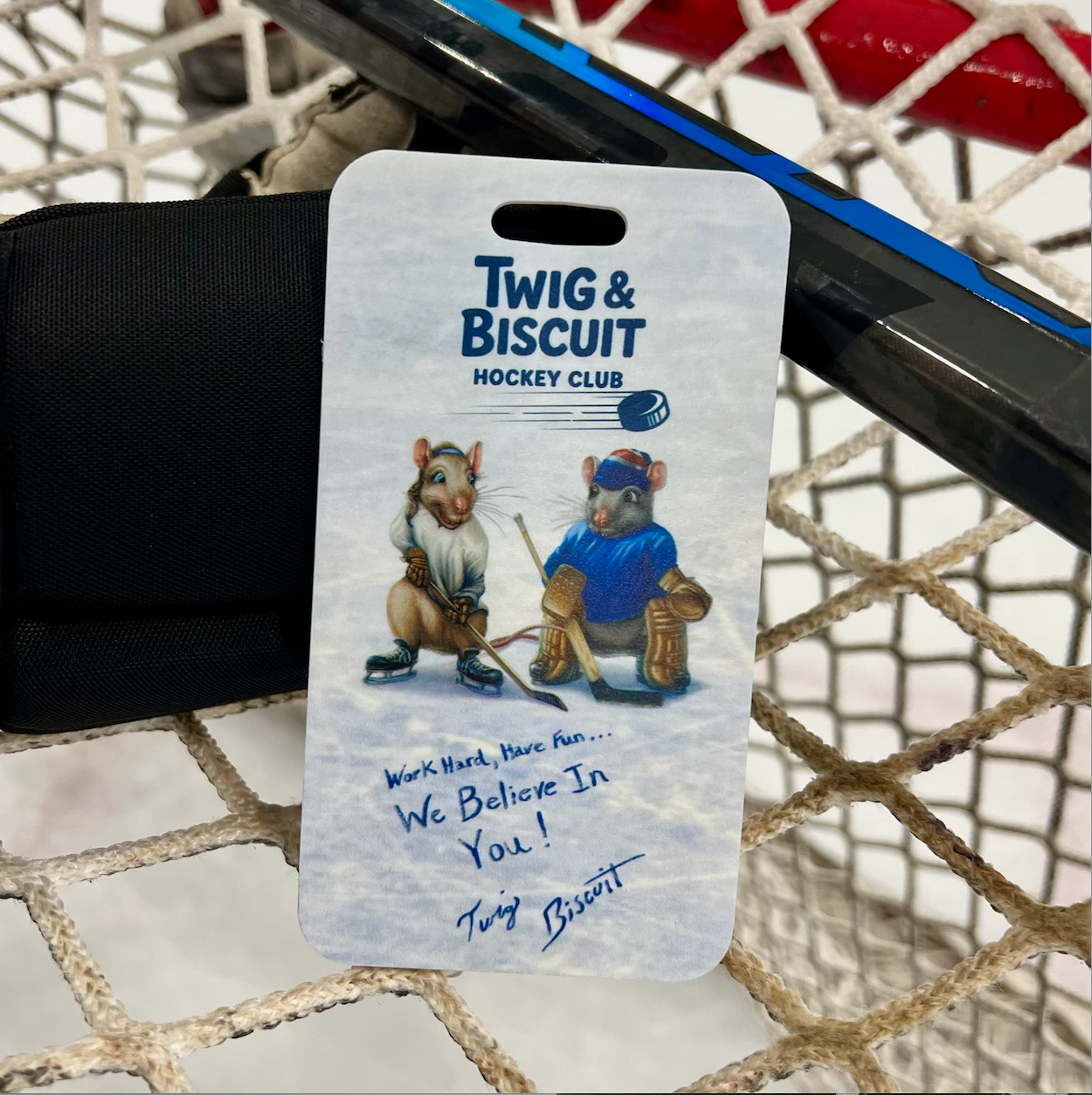 Hockey-themed luggage tag with cartoon bears and text, placed on a hockey stick against a net background.